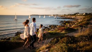 Famille marchant sur un sentier côtier du golfe du Morbihan, avec vue sur la mer et des voiliers au loin.